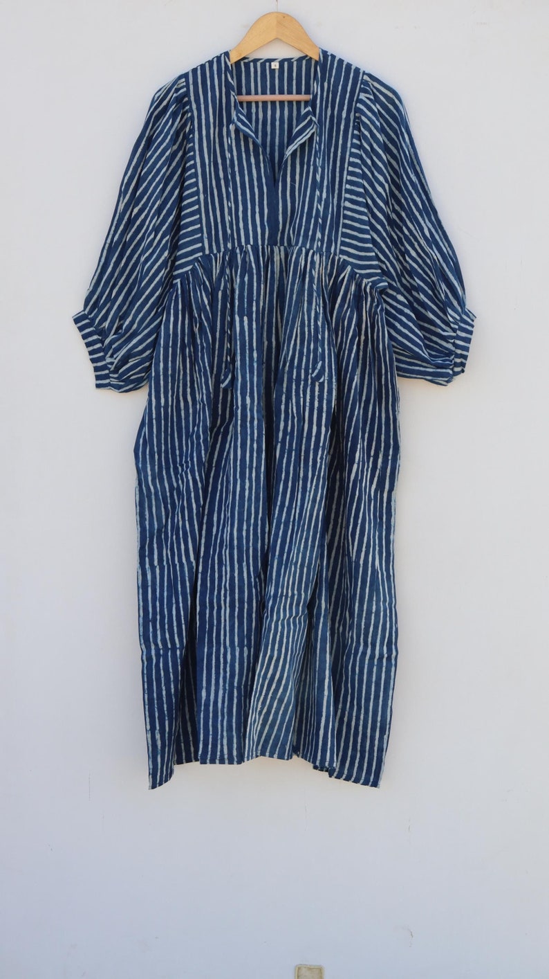 May include: A midi-length dress in deep indigo blue with a white vertical striped pattern. The garment features three-quarter length sleeves, a gathered waistline, and a split V-neckline with a small collar, hanging on a wooden hanger.