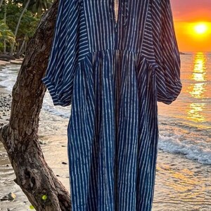 May include: A midi-length dress featuring a dark blue and white vertical striped pattern. The garment has a round neckline with a small front tie closure, three-quarter length puff sleeves with cuffed hems, and a gathered waistline.