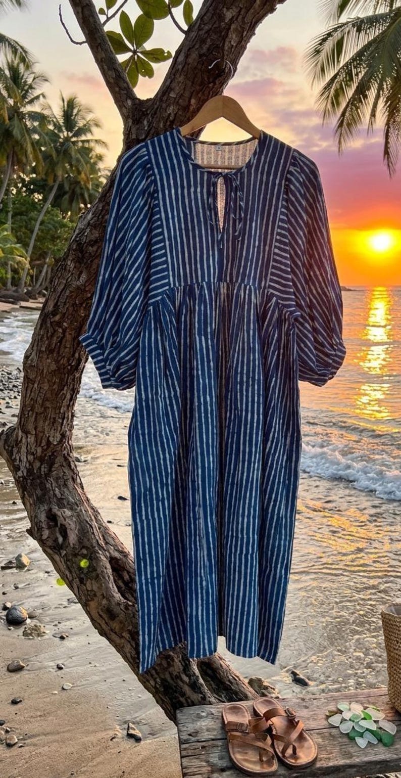 May include: A midi-length dress featuring a dark blue and white vertical striped pattern. The garment has a round neckline with a small front tie closure, three-quarter length puff sleeves with cuffed hems, and a gathered waistline.