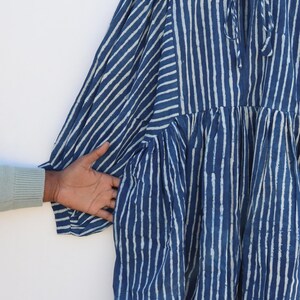 May include: A knee-length dress featuring a dark blue and white vertical striped pattern. The garment has long, gathered sleeves, a round neckline with a tie closure, and a loose, flowing silhouette. The fabric displays a textured, hand-dyed appearance.