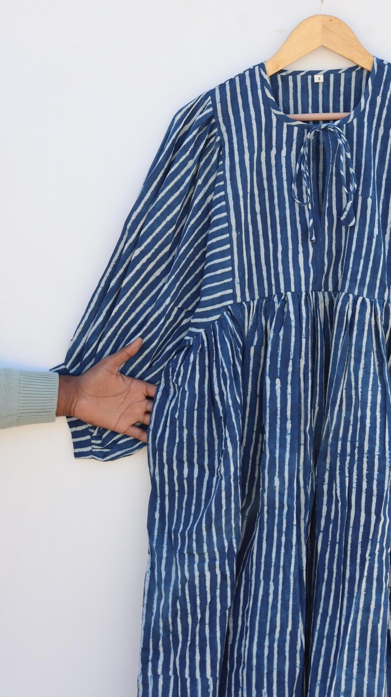 May include: A knee-length dress featuring a dark blue and white vertical striped pattern. The garment has long, gathered sleeves, a round neckline with a tie closure, and a loose, flowing silhouette. The fabric displays a textured, hand-dyed appearance.