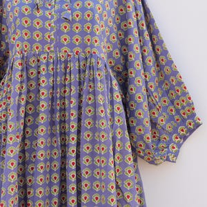 Puede incluir: Un vestido de algod&oacute;n morado con estampado floral en tonos verdes y rojos. El vestido tiene un escote redondo y un lazo en el cuello. Las mangas son 3/4 y tienen un pu&ntilde;o fruncido. El vestido tiene una falda fruncida.