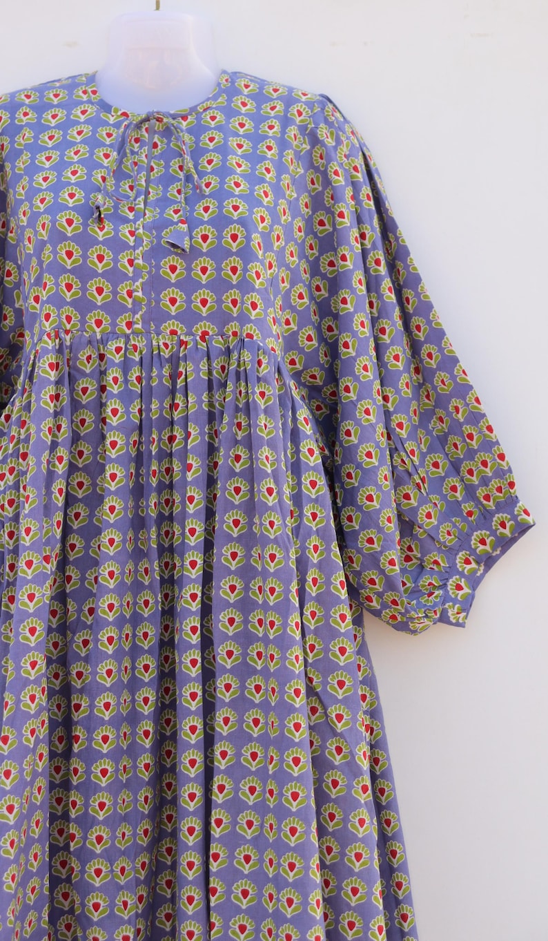 Puede incluir: Un vestido de algod&oacute;n morado con estampado floral en tonos verdes y rojos. El vestido tiene un escote redondo y un lazo en el cuello. Las mangas son 3/4 y tienen un pu&ntilde;o fruncido. El vestido tiene una falda fruncida.