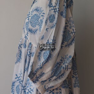 May include: White cotton dress with a blue floral print. The dress has a gathered skirt and a round neckline. The dress is on a white mannequin.
