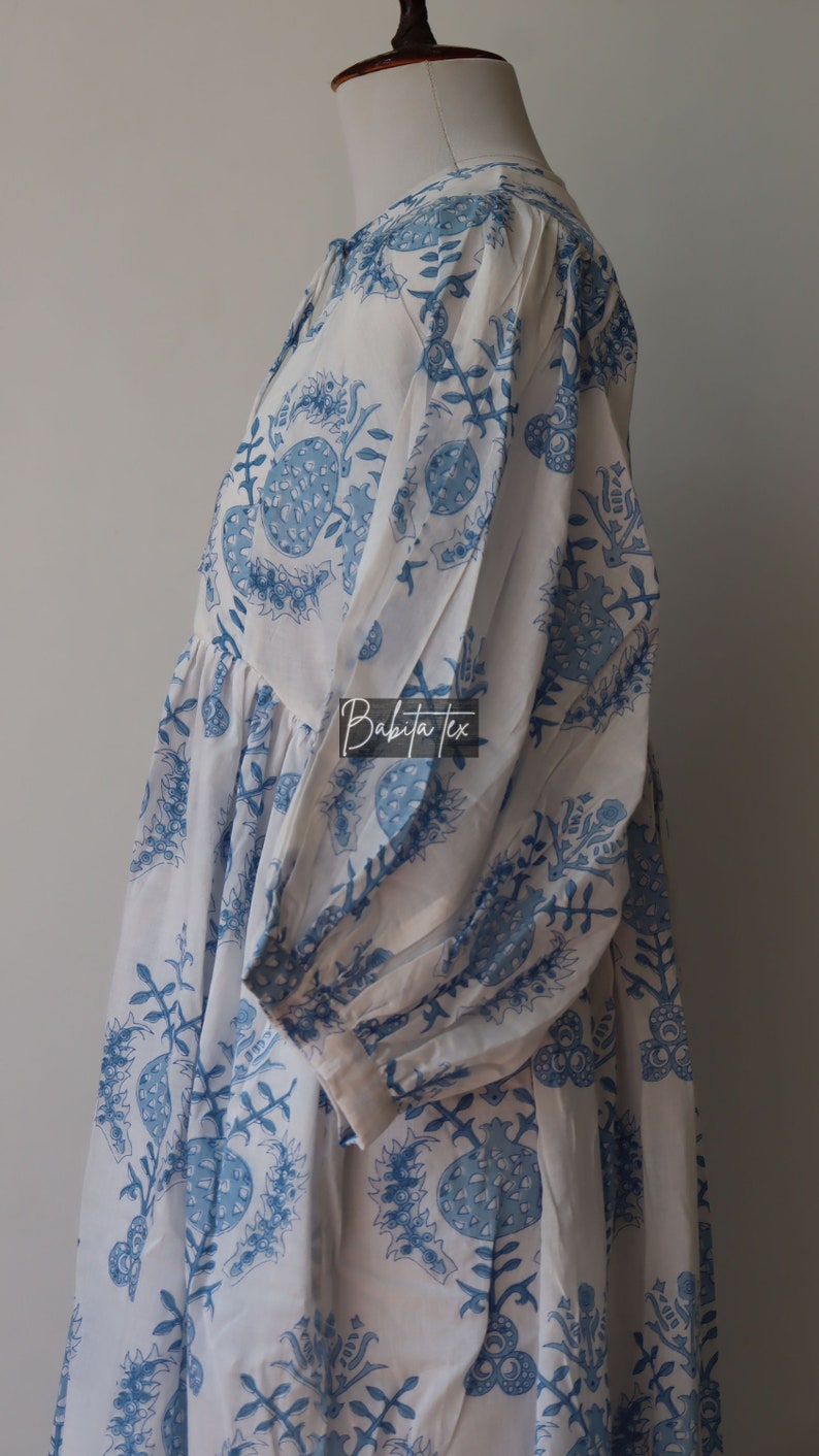 May include: White cotton dress with a blue floral print. The dress has a gathered skirt and a round neckline. The dress is on a white mannequin.