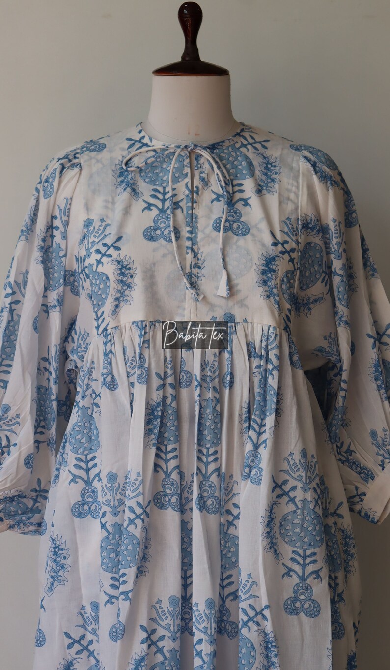 May include: White cotton dress with a blue floral print and a tie at the neck. The dress has a gathered waist and 3/4 sleeves. The dress is made by Babita Tex.