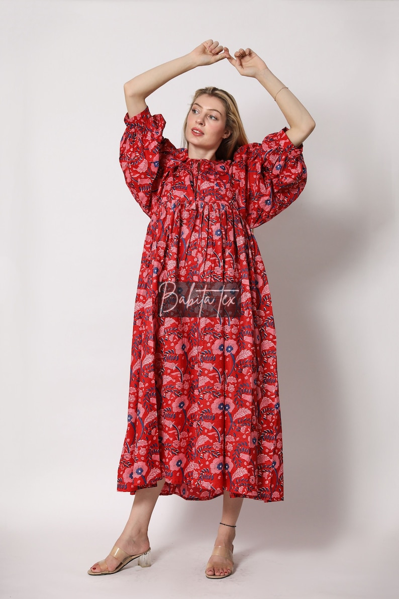 Puede incluir: Un vestido maxi rojo con estampado floral y cintura fruncida. El vestido tiene un escote redondo y mangas 3/4. La modelo lleva sandalias de tac&oacute;n transparentes.