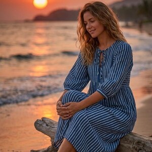 May include: A blue and white striped midi dress featuring three-quarter length sleeves, a gathered waist, and a round neckline with a tie-front closure. The garment is constructed with a tiered skirt design and a relaxed, flowing silhouette.