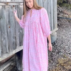 May include: A woman wearing a pink floral dress with a tie at the neck. The dress has a loose fit and is made of a lightweight fabric. She is standing in front of a wooden building.