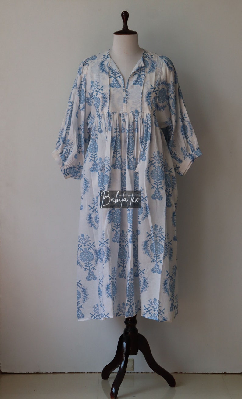 May include: A white cotton dress with a blue floral print. The dress has a V-neckline, 3/4 sleeves, and a gathered waist. The dress is on a mannequin and the text "Babita Tex" is visible on the dress.