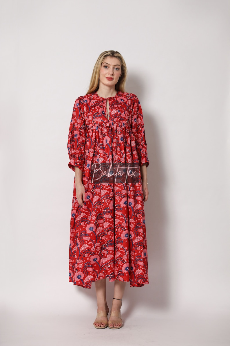 Puede incluir: Un vestido maxi rojo con estampado floral y un lazo en el cuello. El vestido tiene una falda fruncida y mangas 3/4.