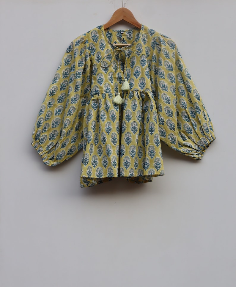 May include: A yellow cotton top with a blue floral print and a tie at the neck. The top has long sleeves with a gathered cuff and a gathered waist.