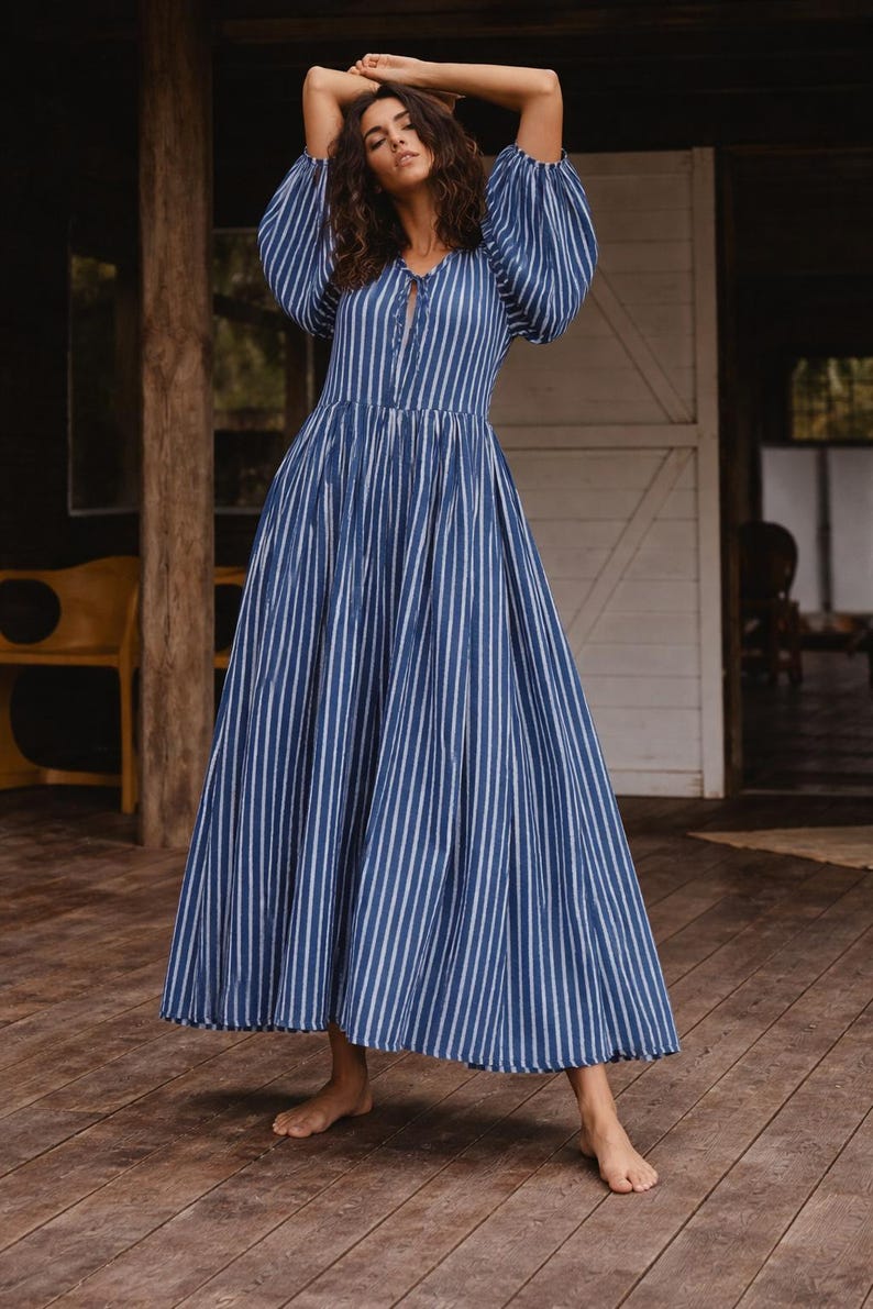 May include: A floor-length dress featuring a blue and white vertical striped pattern, voluminous puff sleeves, and a front tie closure at the neckline. The garment has a fitted bodice that transitions into a full, flowing skirt with a gathered waistline.