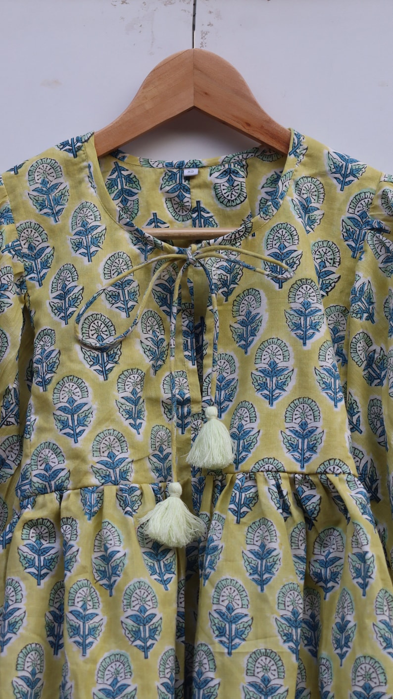 May include: A yellow cotton top with a blue and green floral print. The top has a tie at the neck and tassels at the bottom.