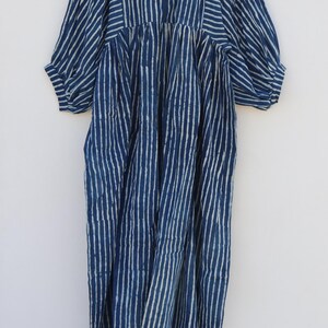 May include: A midi-length dress in deep indigo blue with a white vertical striped pattern. The garment features a round neckline with a tie closure, gathered waist detailing, and voluminous three-quarter length sleeves with elasticated cuffs.