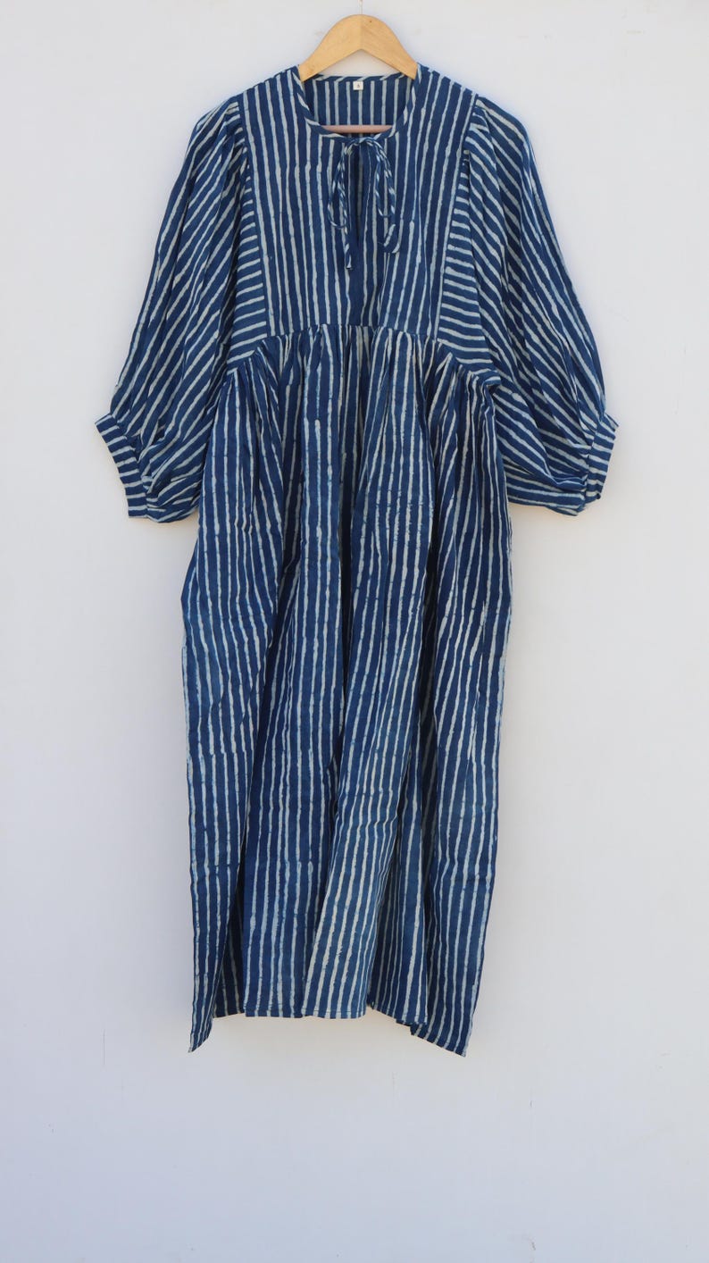May include: A midi-length dress in deep indigo blue with a white vertical striped pattern. The garment features a round neckline with a tie closure, gathered waist detailing, and voluminous three-quarter length sleeves with elasticated cuffs.
