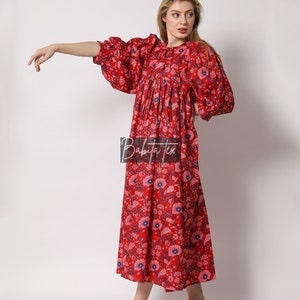 Puede incluir: Un vestido maxi rojo con estampado floral y mangas abullonadas. El vestido tiene una cintura fruncida y un ajuste holgado. La modelo lleva el vestido con sandalias de tac&oacute;n transparentes.