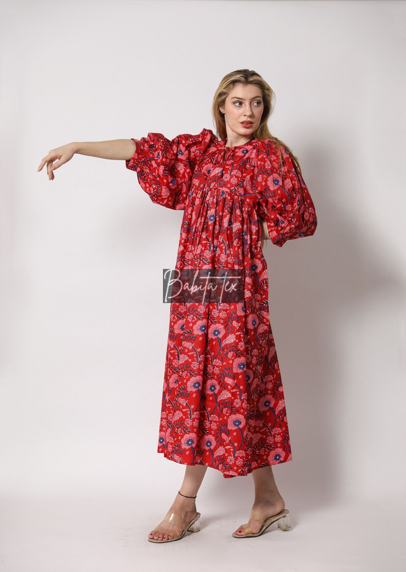 Puede incluir: Un vestido maxi rojo con estampado floral y mangas abullonadas. El vestido tiene una cintura fruncida y un ajuste holgado. La modelo lleva el vestido con sandalias de tac&oacute;n transparentes.