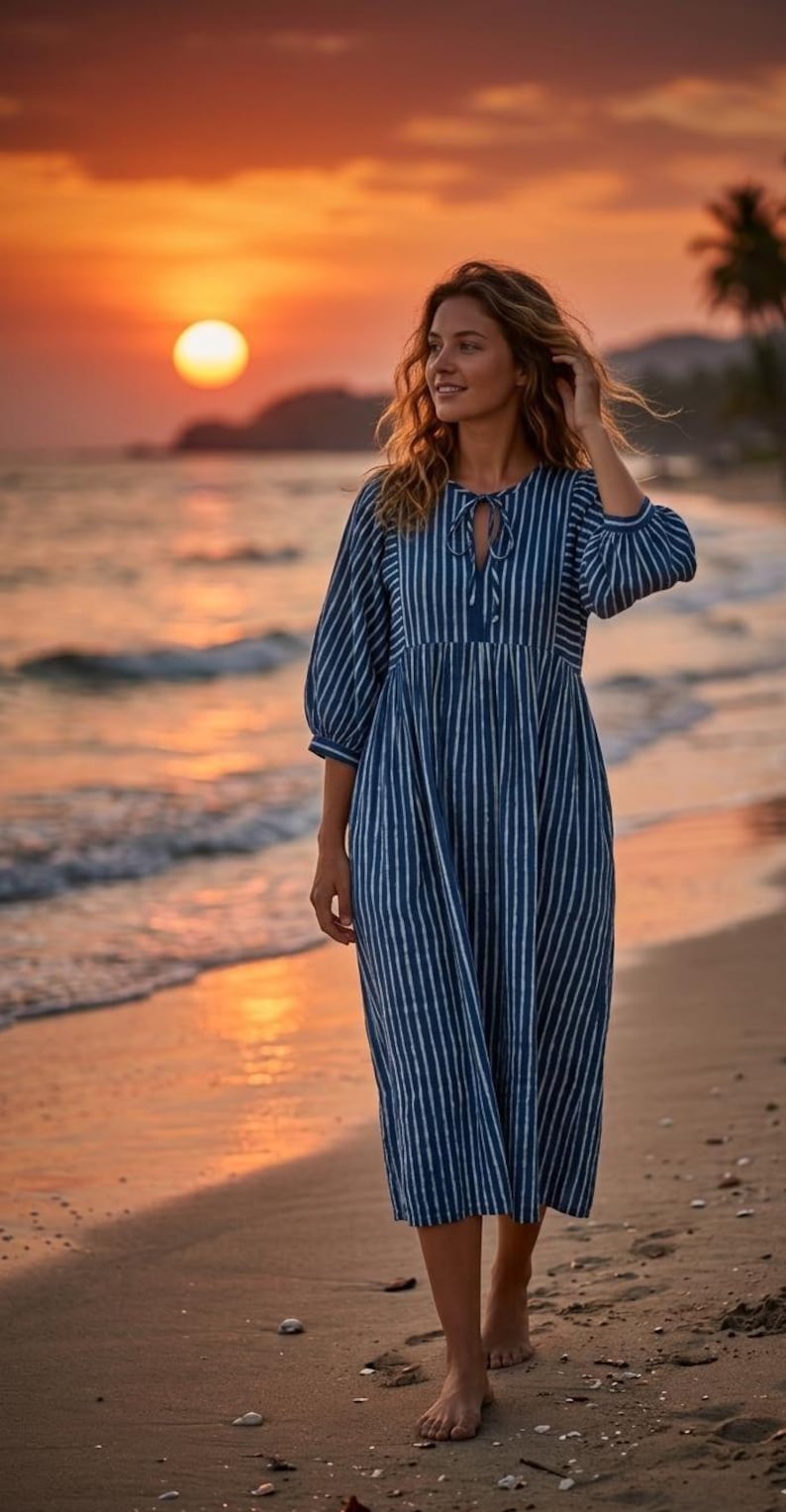 May include: A midi-length dress featuring a dark blue and white vertical striped pattern, three-quarter length sleeves with gathered cuffs, and a round neckline with a central tie closure. The garment has a relaxed fit with a gathered waistline.