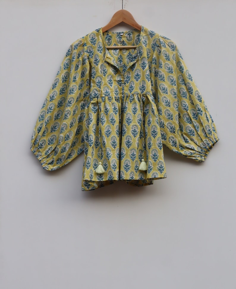 May include: A yellow cotton top with a blue floral print and long sleeves. The top has a V-neck and a gathered waist. The sleeves are gathered at the cuffs and have a tassel detail.