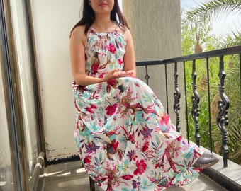 May include: A woman wearing a white maxi dress with a floral print. The dress has a halter neckline and a flowing skirt. She is sitting on a chair on a balcony.