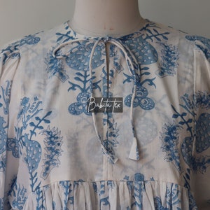 May include: A white cotton tunic with a blue floral print and a tie at the neck. The tunic has a gathered skirt and puffed sleeves. The text "Babita Tex" is visible on the front of the tunic.