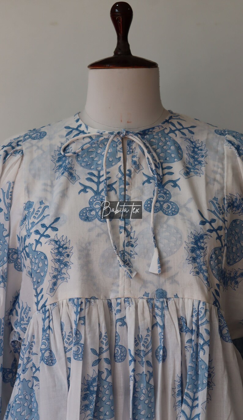 May include: A white cotton tunic with a blue floral print and a tie at the neck. The tunic has a gathered skirt and puffed sleeves. The text "Babita Tex" is visible on the front of the tunic.