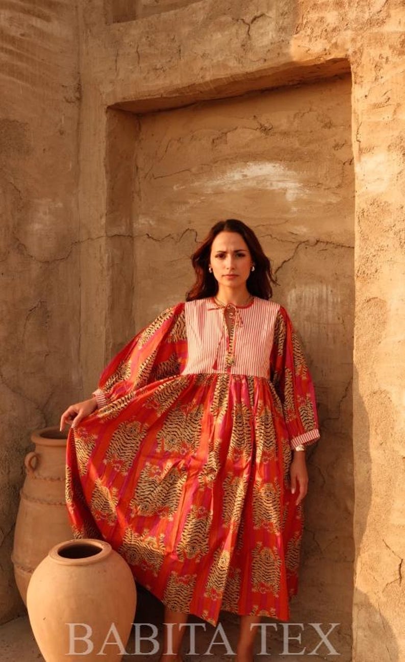 Puede incluir: Un vestido largo y fluido de color rojo con estampado de tigre y corpi&ntilde;o a rayas. El vestido tiene mangas largas y lo lleva una persona. El texto "BABITA TEX" es visible en la parte inferior de la imagen. Es una prenda vibrante.
