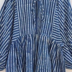 May include: A blue and white striped tunic featuring a round neckline with a tie closure, long puff sleeves, and a gathered waistline. The fabric displays a vertical stripe pattern on the bodice and sleeves, with a size S tag at the collar.