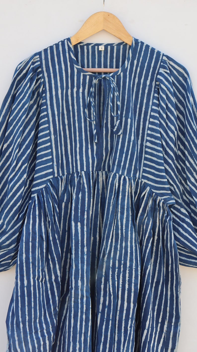 May include: A blue and white striped tunic featuring a round neckline with a tie closure, long puff sleeves, and a gathered waistline. The fabric displays a vertical stripe pattern on the bodice and sleeves, with a size S tag at the collar.