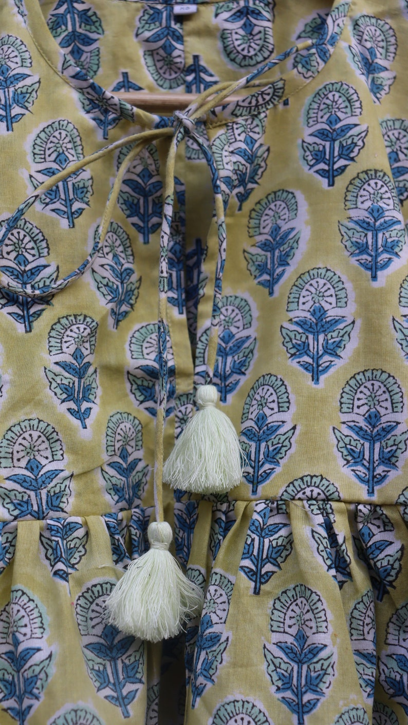 May include: A yellow cotton top with a blue and green floral print. The top has a tie at the neck and tassels at the bottom.