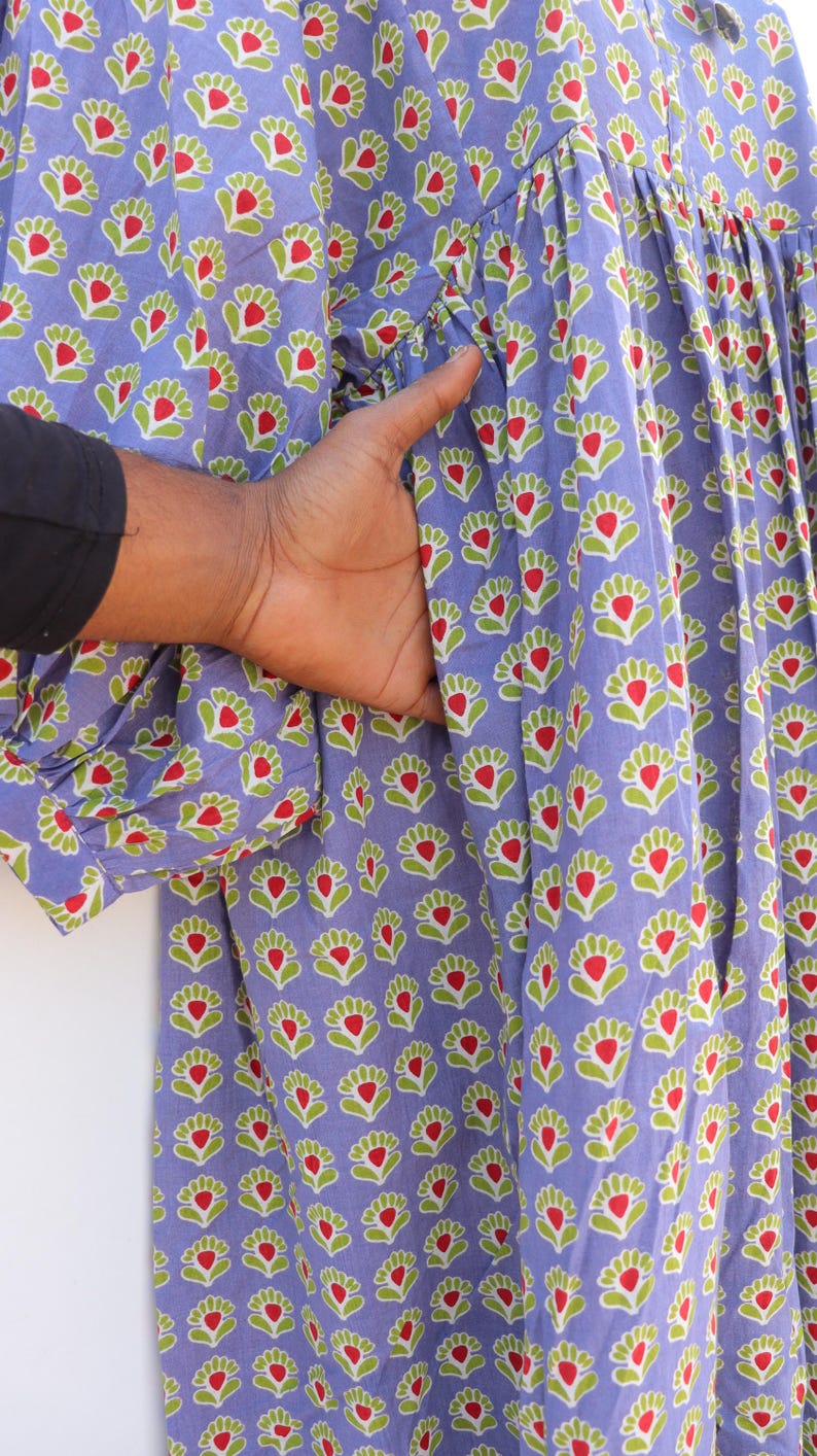 Puede incluir: Un vestido largo de algod&oacute;n azul con estampado floral con flores rojas y verdes. El vestido tiene una falda fruncida y un frente abotonado.