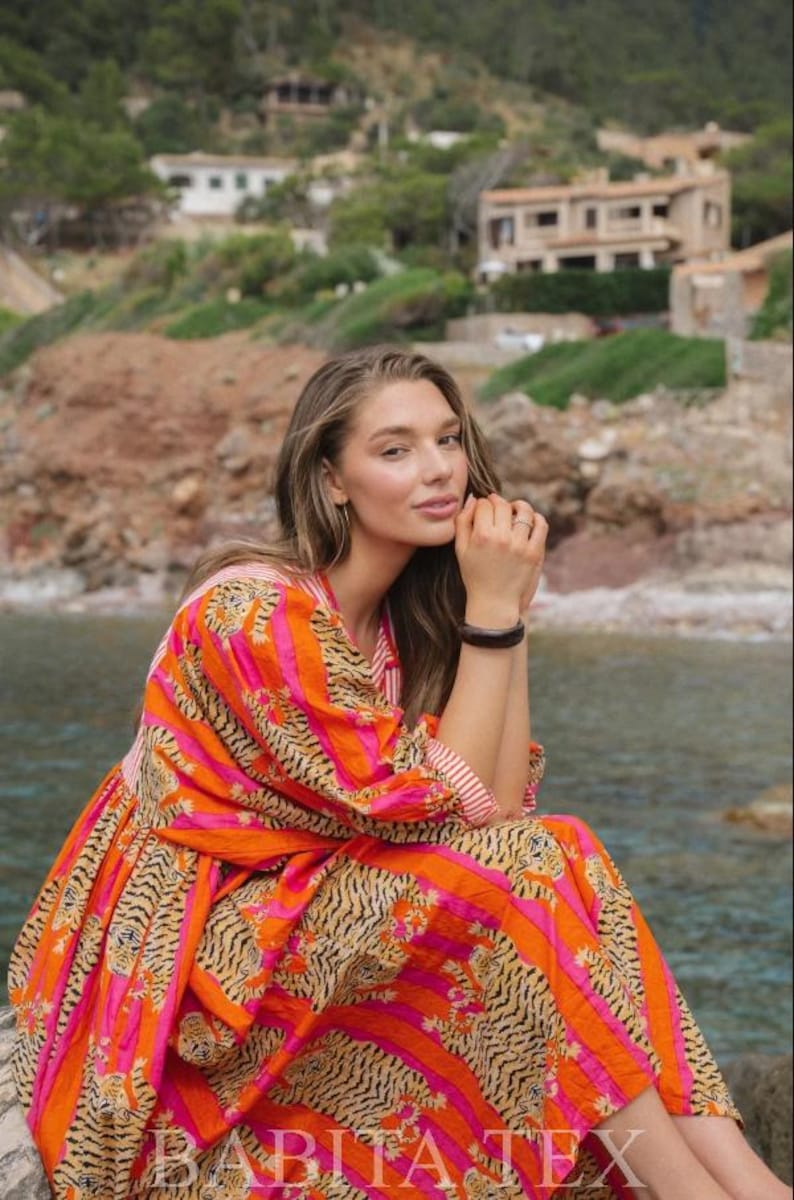 Puede incluir: Vestido vibrante con estampado de rayas de tigre y rayas verticales en naranja y rosa. El vestido tiene mangas largas y una falda fluida. El texto "BABITA TEX" es visible en la parte inferior del vestido.