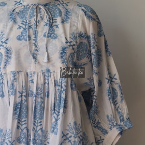 May include: White cotton tunic with a blue floral print. The tunic has a gathered waist and a tie at the neck. The text "Babita Tex" is visible on the front of the tunic.