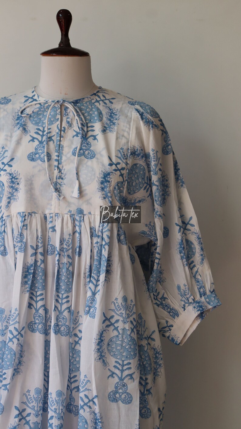 May include: White cotton tunic with a blue floral print. The tunic has a gathered waist and a tie at the neck. The text "Babita Tex" is visible on the front of the tunic.