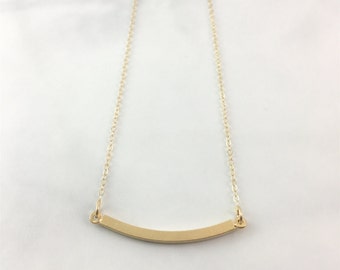 Puede incluir: Un collar de oro con un colgante de barra curvada. La cadena es de estilo delicado, de eslabones finos.