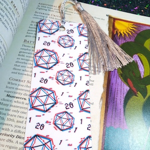 May include: A white bookmark with a glittery tassel. The bookmark is covered in a repeating pattern of 20-sided dice with a glitch effect in red and blue.