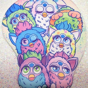 May include: A mouse mat with a design of multiple Furby characters in shades of blue, pink, green, and yellow. The background is light-coloured with a glittery effect. The Furby characters are stylized.