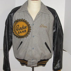 May include: A grey and black varsity jacket with a gold and black "Purdue Band" patch on the left side and the name "Steve" embroidered on the right side. The jacket has black leather sleeves with gold and black striped cuffs and waistband.