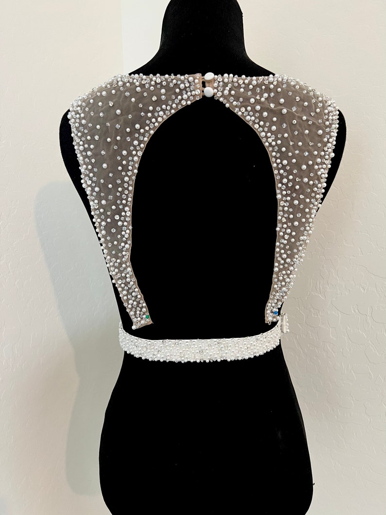 May include: A white beaded bodice with a low back and a beaded belt. The bodice is made of a sheer fabric with small white beads sewn on.