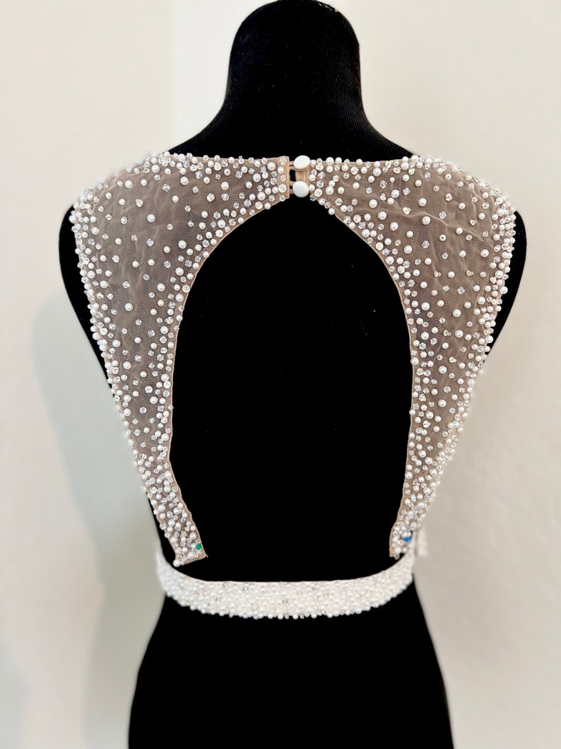 May include: A white beaded bodice with a keyhole back and a beaded belt. The bodice is made of a sheer fabric with a delicate pattern.