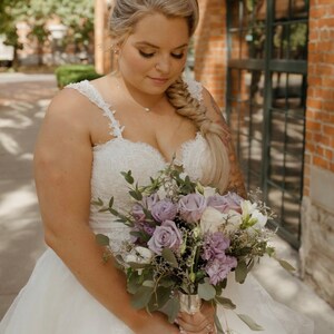 May include: A bride in a white lace wedding dress holds a bouquet of purple and white flowers. She is standing in front of a brick building with a green door.