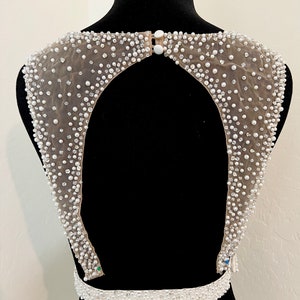 May include: A white beaded bodice with a low back and a beaded belt. The bodice is made of a sheer fabric with small white beads and pearls.