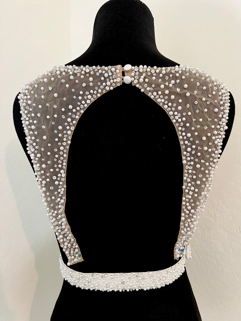 May include: A white beaded bodice with a low back and a beaded belt. The bodice is made of a sheer fabric with small white beads and pearls.