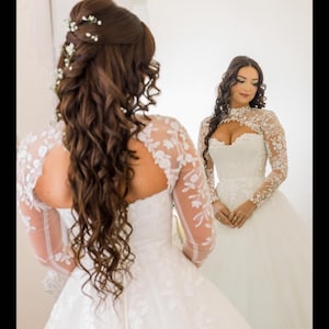 May include: A bride in a white wedding dress with lace sleeves and bodice, reflected in a mirror. Her long, wavy brown hair is styled with small white flowers. The dress features floral lace details.