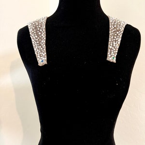May include: A pair of beaded shoulder straps with a light brown fabric base. The straps are adorned with small, white beads and rhinestones.