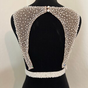 May include: A white beaded bodice with a low back and a beaded belt. The bodice is made of a sheer fabric with small white beads and sequins.