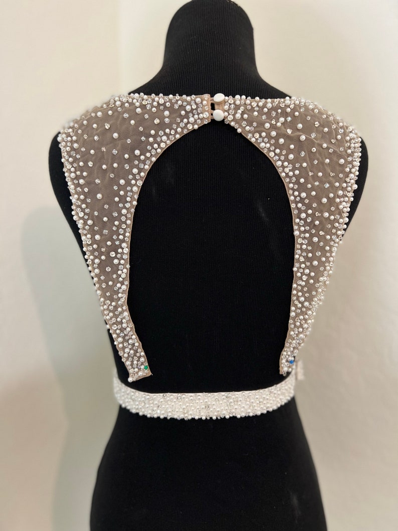 May include: A white beaded bodice with a low back and a beaded belt. The bodice is made of a sheer fabric with small white beads and sequins.