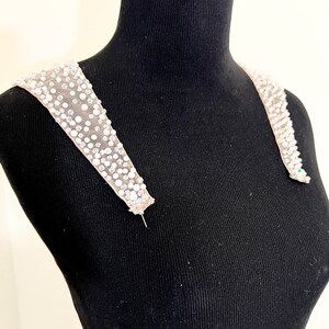 May include: A pair of white beaded shoulder straps for a dress or top. The straps are made of a sheer fabric with small white beads sewn on.