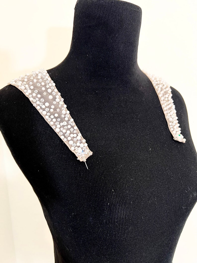 May include: A pair of white beaded shoulder straps for a dress or top. The straps are made of a sheer fabric with small white beads sewn on.